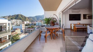 A balcony with a dining table and chairs, a wicker chair, and a view of a cityscape and hills. - Top Floor Condo with City View, Rooftop Pool, Gym in Old Town (Puerto Vallarta)