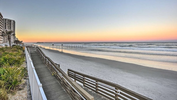 Een strand met een houten promenade, een pier die in de oceaan uitsteekt, en gebouwen op de achtergrond.
