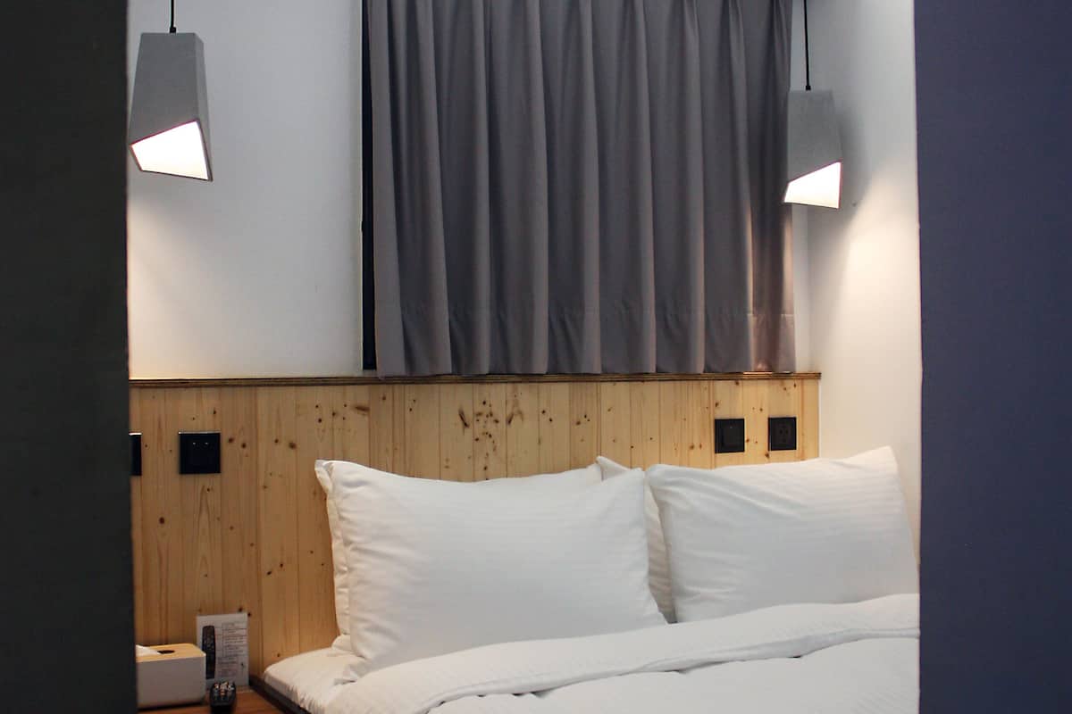 A modern hotel room with a wooden headboard, a large bed with white linens, two bedside tables, and two wall-mounted lamps.