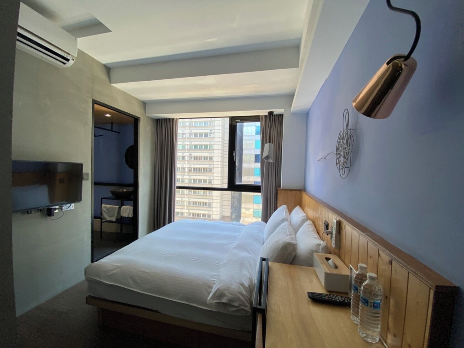 Une chambre d’hôtel avec un lit, un bureau doté d’une lampe, une télévision et une fenêtre donnant sur un immeuble.