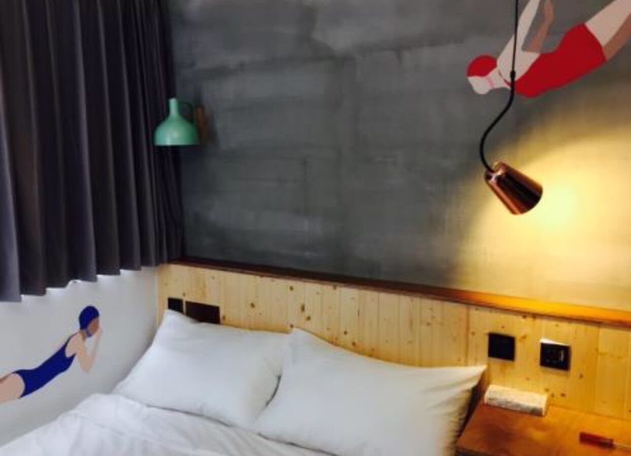 A hotel room with a wooden headboard, white bedding, a teal wall lamp, and a red and white wall decoration.