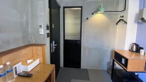 A compact hotel room with a kitchenette, a wooden desk with a remote control, water bottles, a tissue box, and a black door. - Just Inn - City Hall (Taipei)