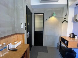 Een kleine hotelkamer met een kitchenette, een houten bureau met een afstandsbediening, waterflessen, een zakdoosje en een zwarte deur.