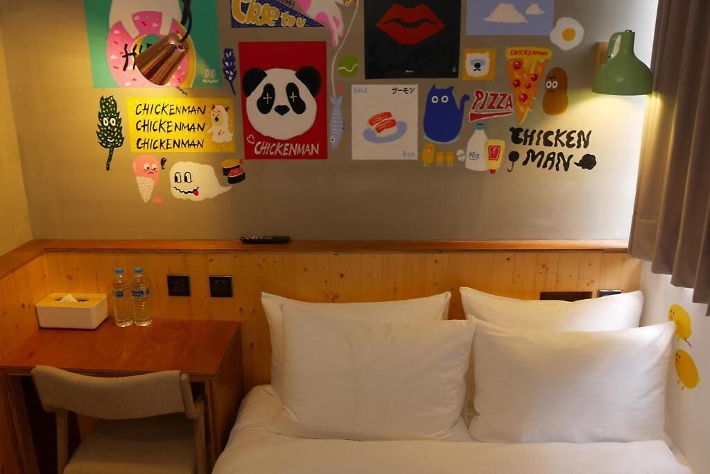 A hotel room with a wooden headboard, a small table with two bottles of water, a tissue box, and a wall decorated with various cartoon characters and words like "CHICKENMAN" and "PIZZA".