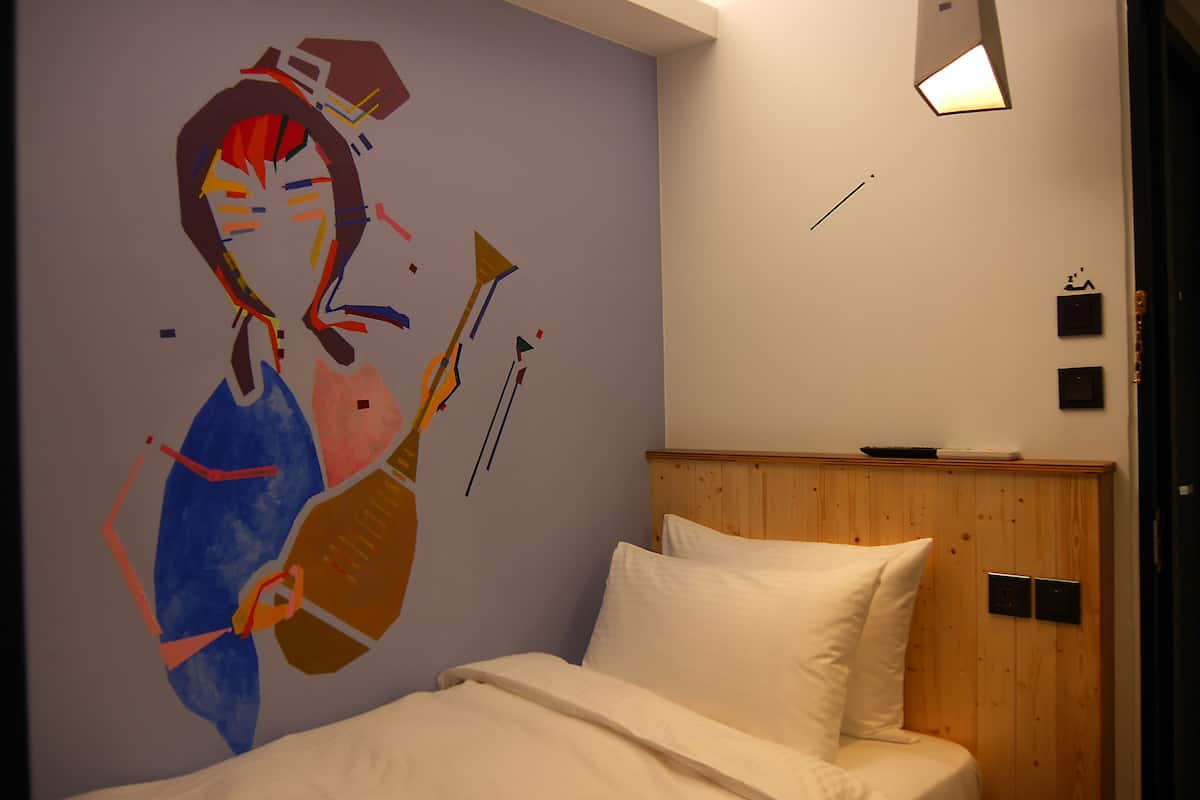 A bedroom with a bed, a wooden headboard, a wall mural, and a wall-mounted light fixture.