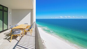 A balcony with a wooden table and chairs overlooking a beach with clear turquoise water and a distant coastline. - The Oasis at Orange Beach, formerly known as Phoenix West II, Unit 2310 (Orange Beach)