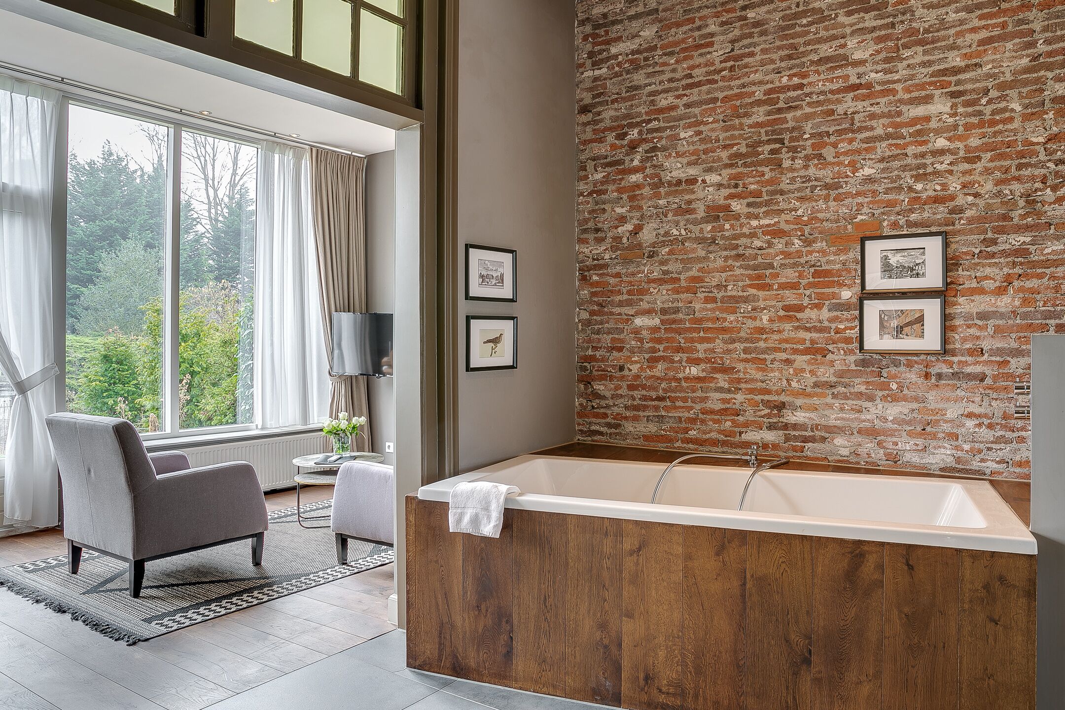 A modern bathroom with a large bathtub, wooden paneling, and a brick wall. There are two framed pictures on the wall and a window with curtains.