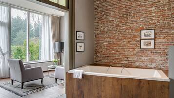 A modern bathroom with a large bathtub, wooden paneling, and a brick wall. There are two framed pictures on the wall and a window with curtains.
