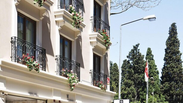 A multi-story hotel with balconies adorned with flowers, a clear sky, and a flag in the background.