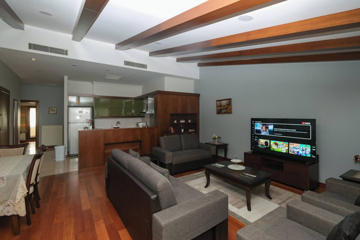 A modern living room with a sofa, coffee table, and TV. There is a kitchen in the background with wooden cabinets and a refrigerator.