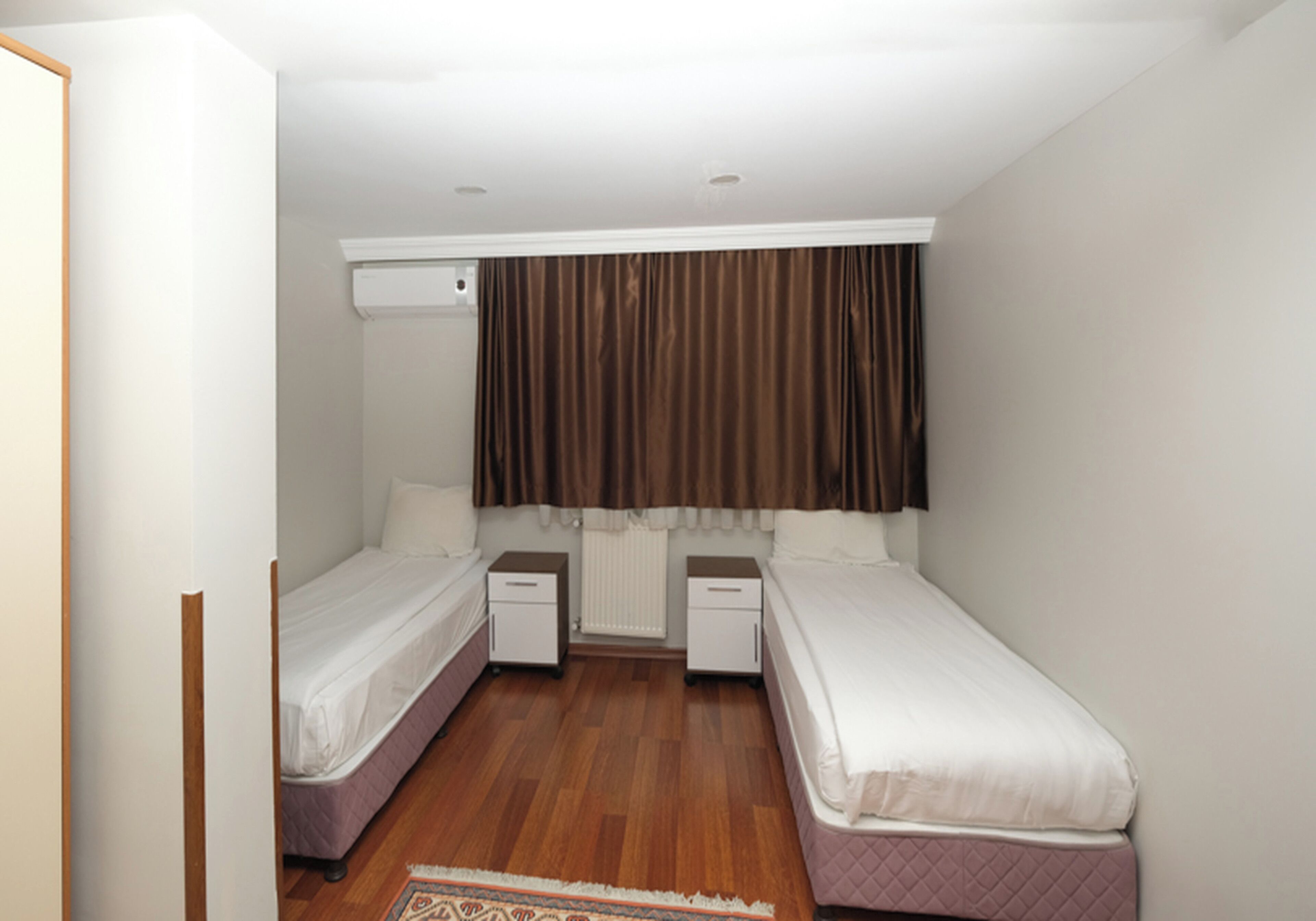 Una habitación pequeña y limpia con dos camas individuales, un radiador blanco y una cortina marrón.