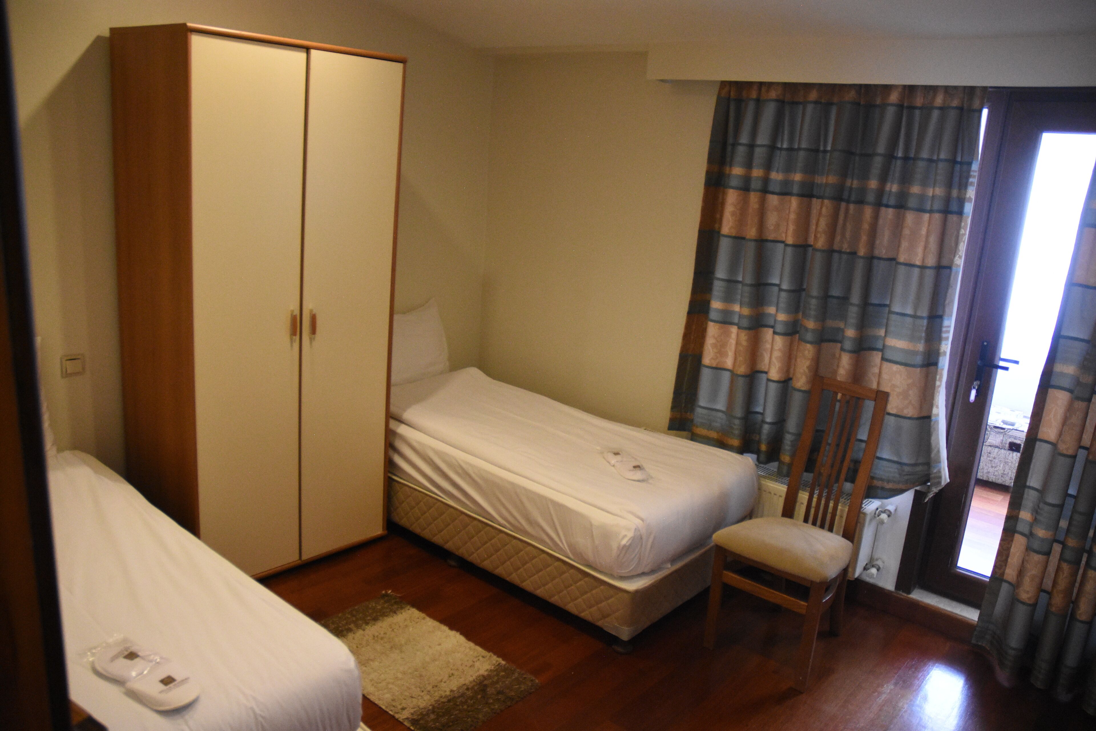 A small, neatly arranged bedroom with a single bed, a wooden wardrobe, a chair, and a window with curtains.