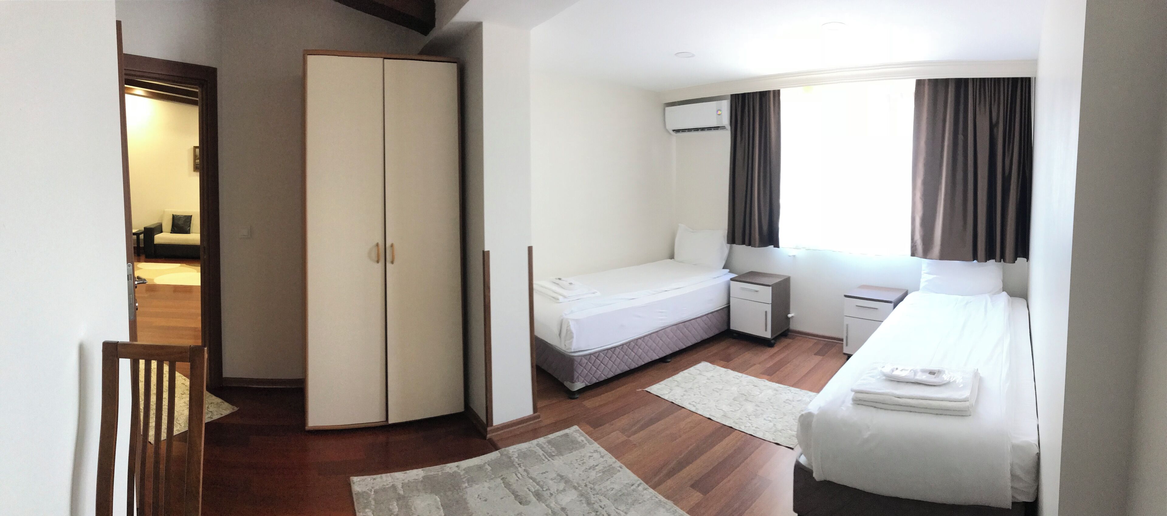 A hotel room with a wardrobe, a single bed, a bedside table, a small desk, and a window with curtains.