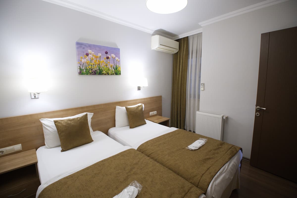 A hotel room with two beds, a painting on the wall, an air conditioning unit, a window with curtains, and a door.