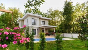 Una casa de dos pisos con piscina, rodeada de un césped cuidado y plantas con flores.