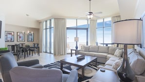 A spacious living room with a large window, a ceiling fan, a sofa set, and a dining area with chairs and a table. - The Oasis at Orange Beach, formerly known as Phoenix West II, Unit 2308 (Orange Beach)