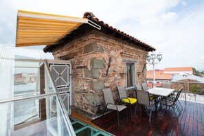 Un edificio rústico de piedra con tejado de tejas, una puerta pequeña y una ventana. Hay una mesa con sillas y un cojín amarillo en una terraza exterior.