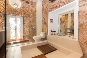 Un baño con paredes de piedra, un inodoro blanco, un espejo grande y un tapete de madera en el suelo.