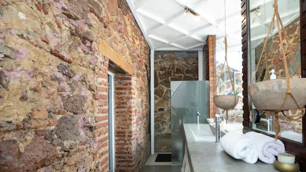 A stone-walled bathroom with a glass shower enclosure, a hanging wooden bath tub, and neatly rolled towels on the floor.