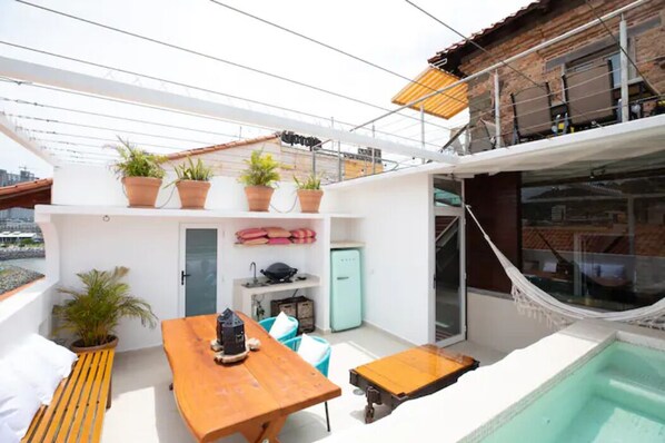 Una terraza en la azotea con mesa y sillas de madera, plantas en macetas, una piscina pequeña y una hamaca.