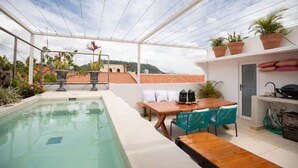 A rooftop pool area with a wooden table and chairs, surrounded by potted plants and a view of buildings in the background.
