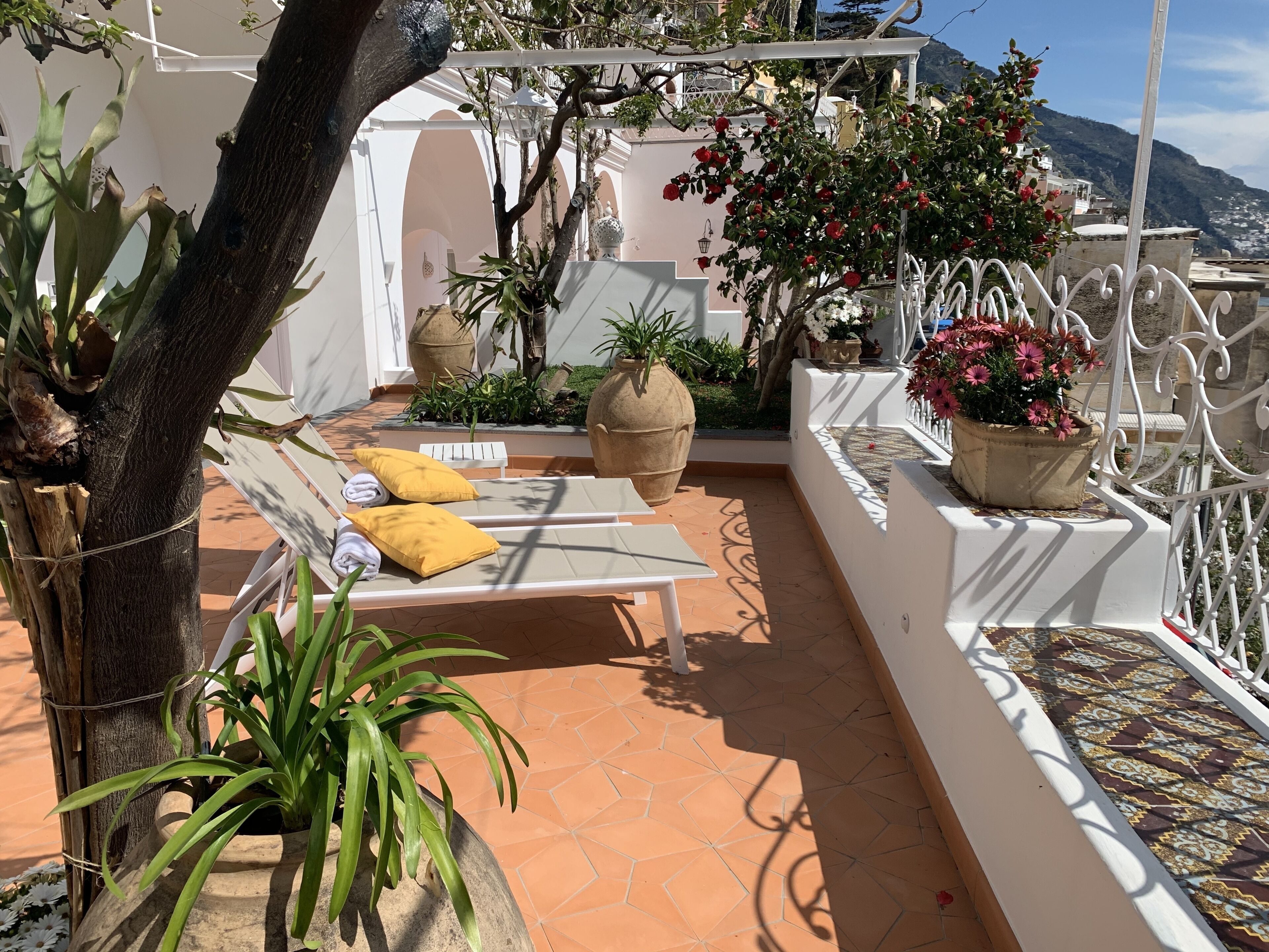 A terrace with a white pergola, a bench with cushions, potted plants, and a view of a mountainous landscape.