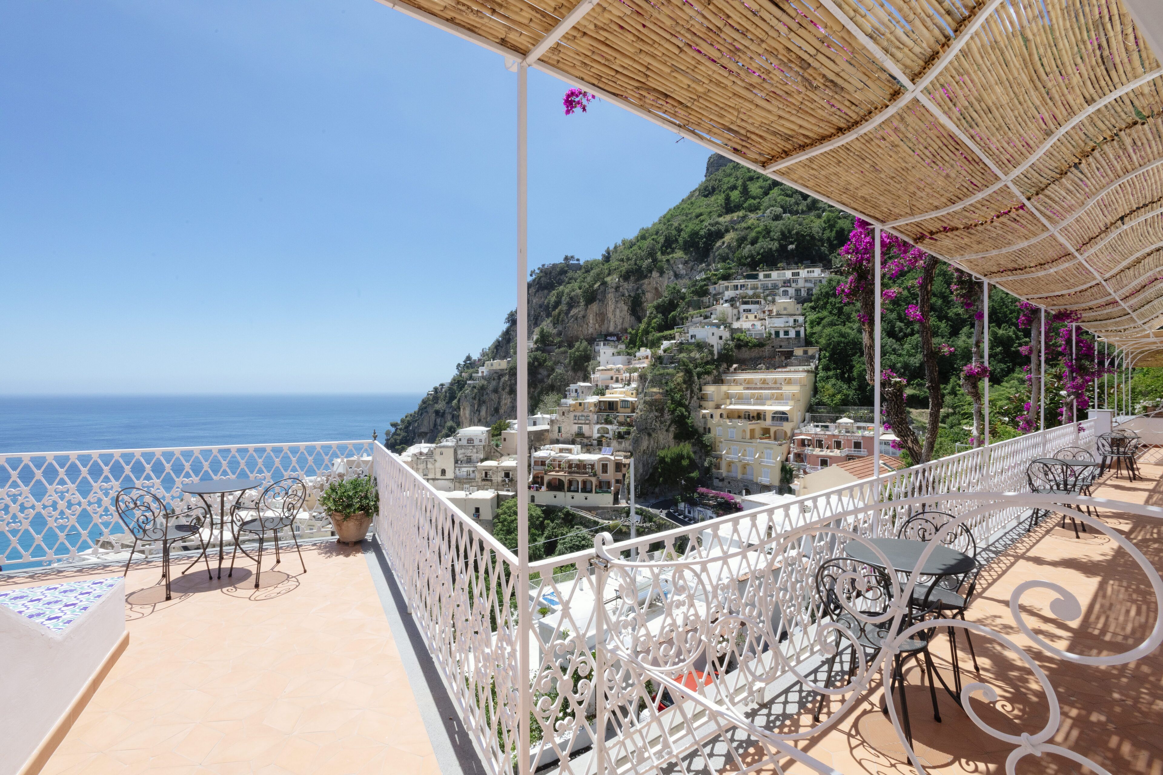 A terrace with outdoor furniture, a view of the sea, and buildings on a hillside.