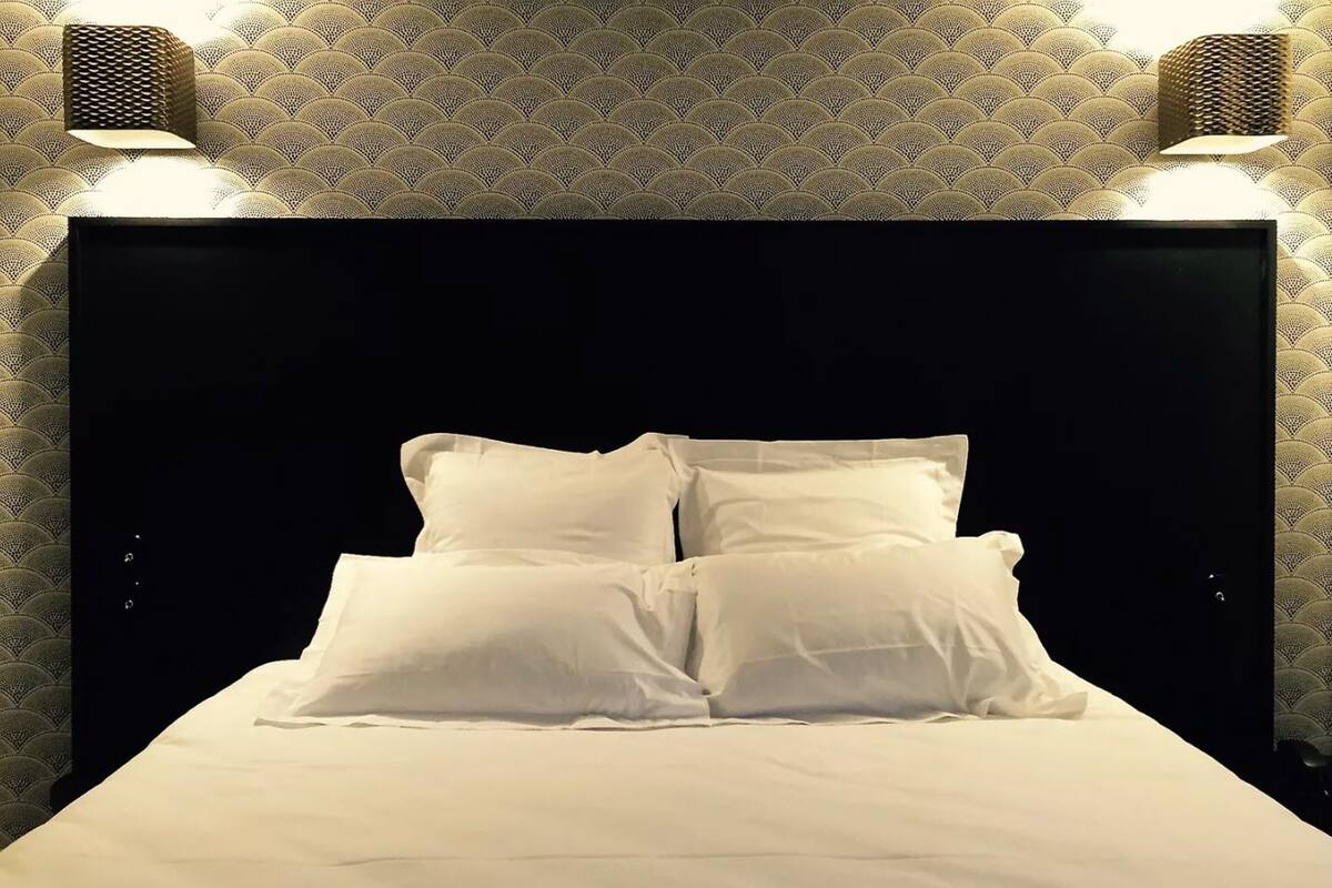 Un lit avec des draps blancs et deux oreillers, placé devant une tête de lit sombre, le tout surmonté d’un papier peint à motifs.
