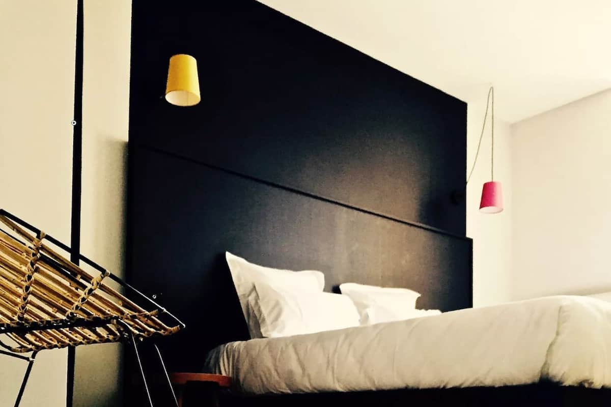 Une chambre à coucher moderne avec une tête de lit sombre, un lit recouvert de linge de lit blanc et deux lampes suspendues.