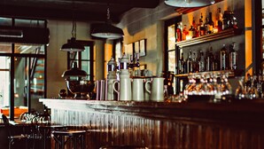 Zona de bar con suelo de madera, taburetes de metal, luces colgantes y estanterías llenas de botellas.