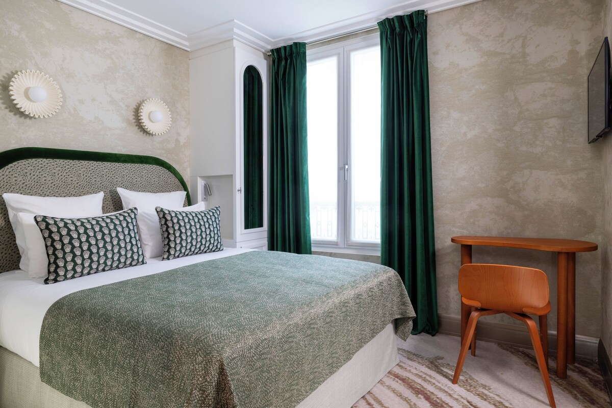 A bedroom with a bed, green curtains, a wooden chair, and a small table.