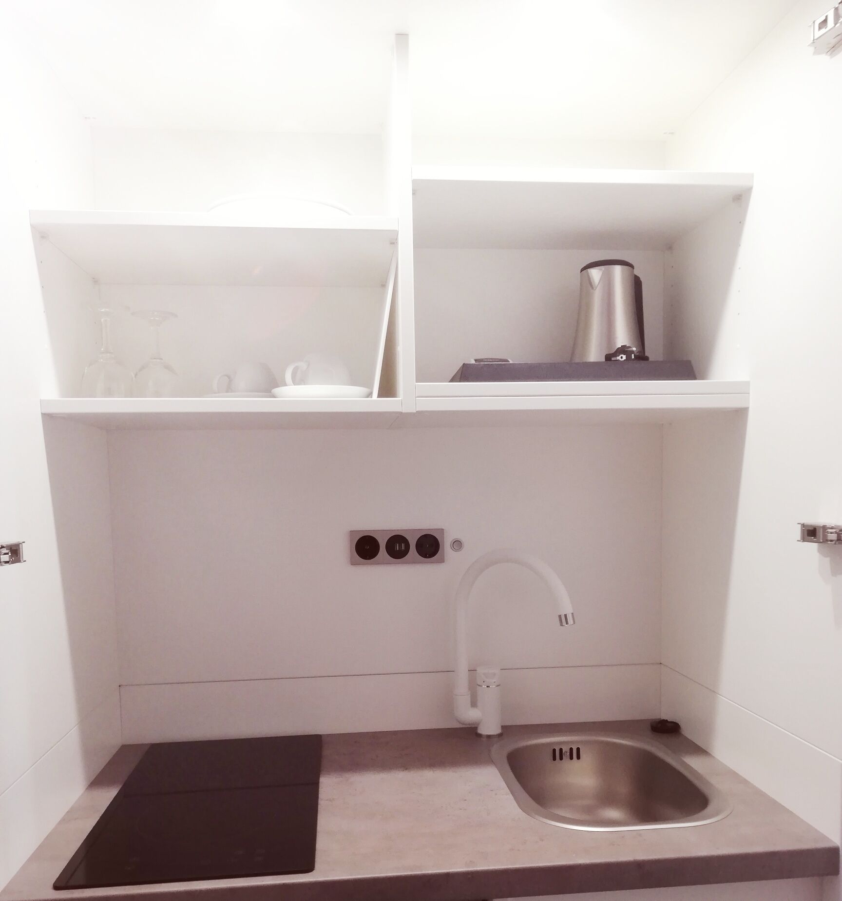 A compact kitchen with a sink, a black mat, and a shelf with kitchenware.