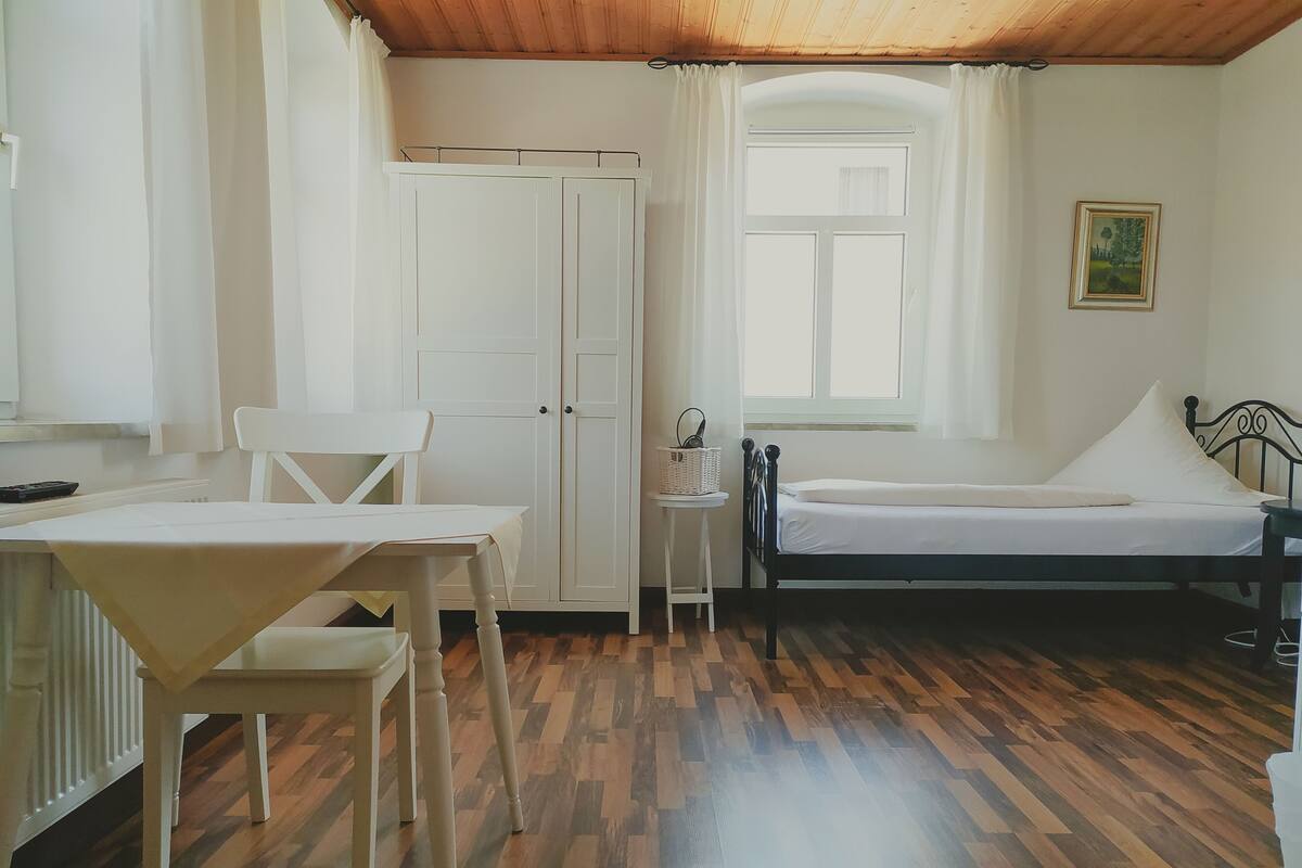 Ett rum med en säng, ett litet bord, en stol, en garderob och ett fönster med gardiner.