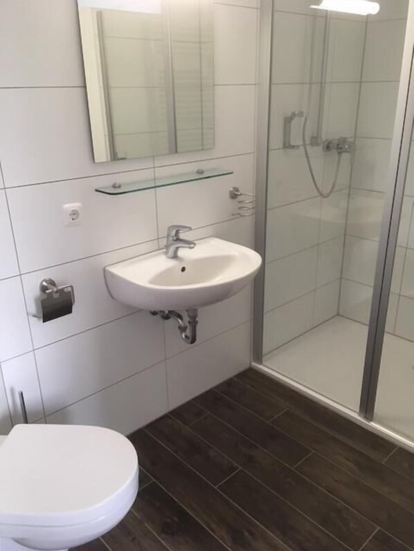 Un baño con lavamanos blanco, espejo, repisa de vidrio, ducha e inodoro.