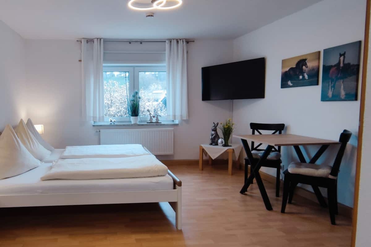 Ett sovrum med en säng, ett skrivbord och en stol. Det finns ett fönster med gardiner, en tv på väggen och en växt på skrivbordet.