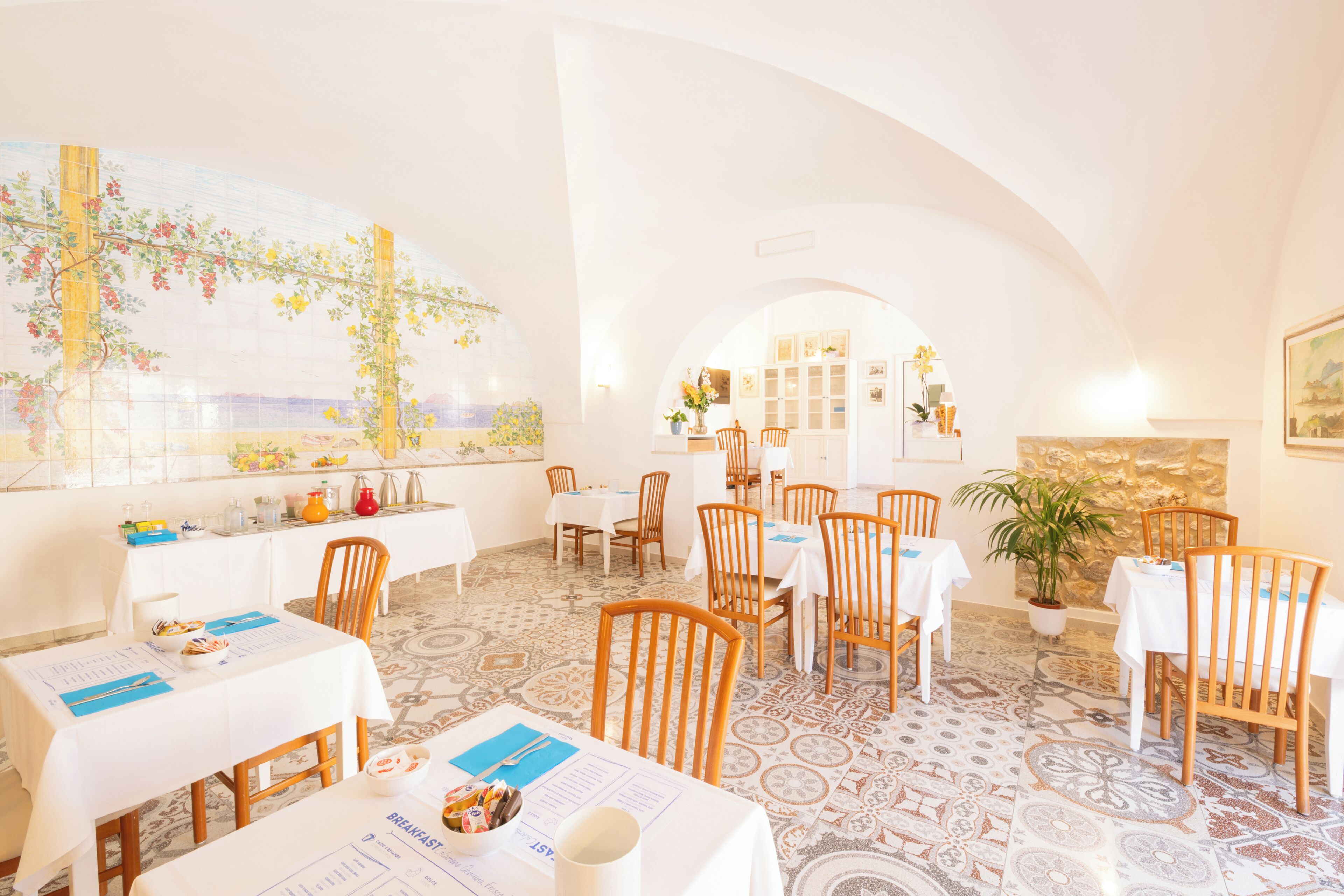 A restaurant with white tables and chairs, a large painting on the wall, and a patterned floor.