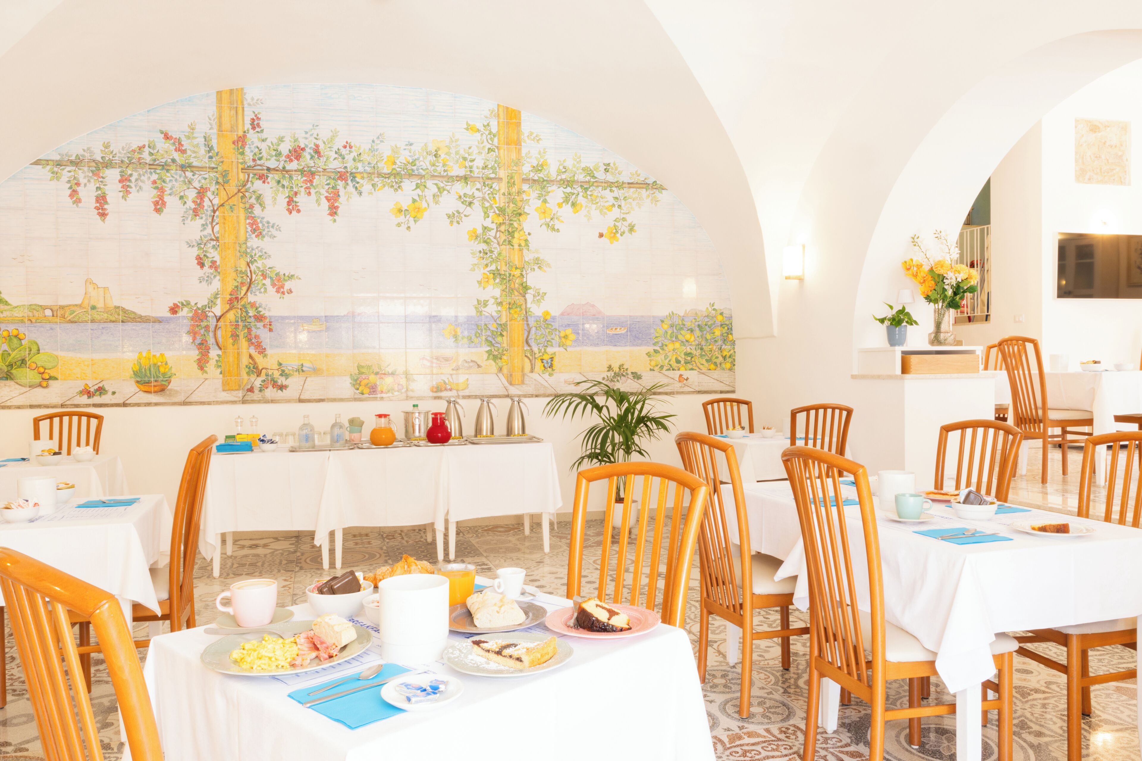 A dining area with tables set for breakfast, a mural on the wall, and a view of the sea.