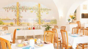 A dining area with tables set for breakfast, a mural on the wall, and a view of the sea.
