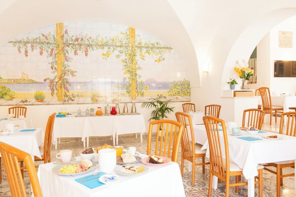 A dining area with tables set for breakfast, a mural on the wall, and a view of the sea.