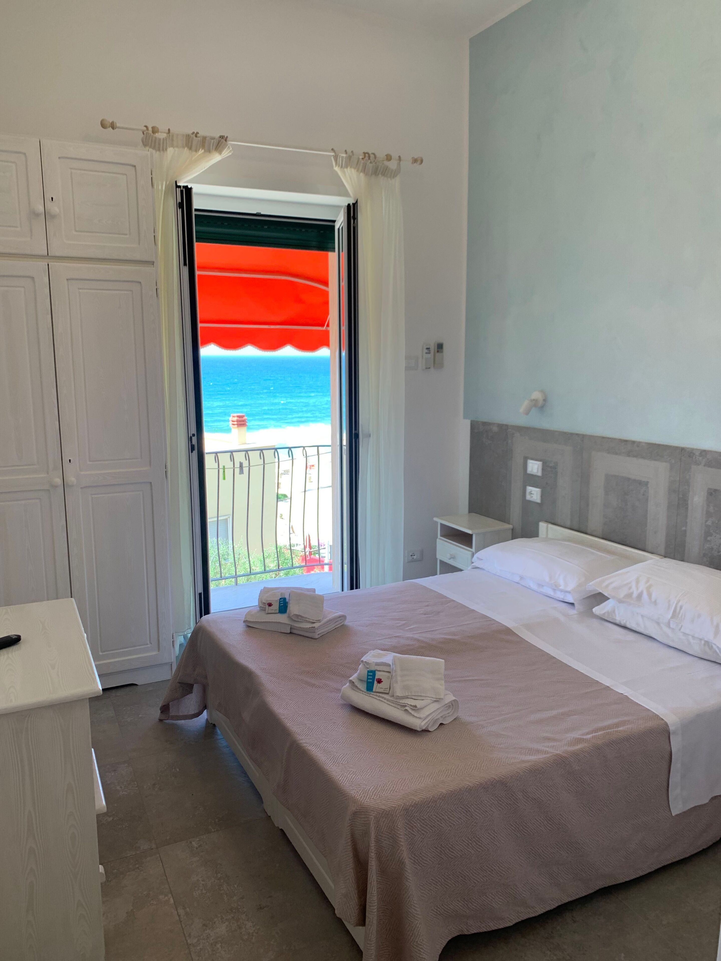A hotel room with a bed, a nightstand, a desk, and a view of the ocean through a balcony door.