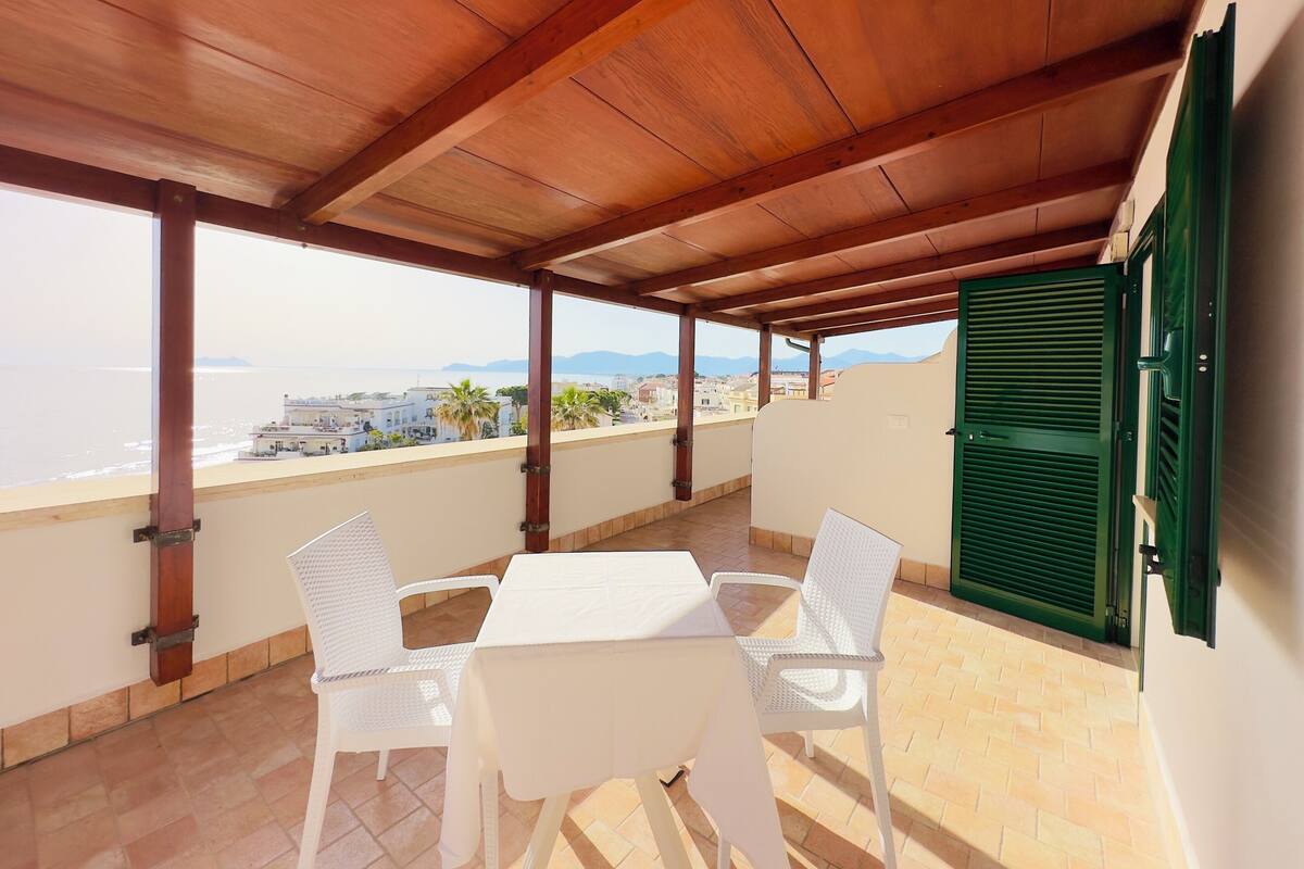 Un espacio al aire libre techado con una mesa y sillas, vista al mar y un edificio al fondo.