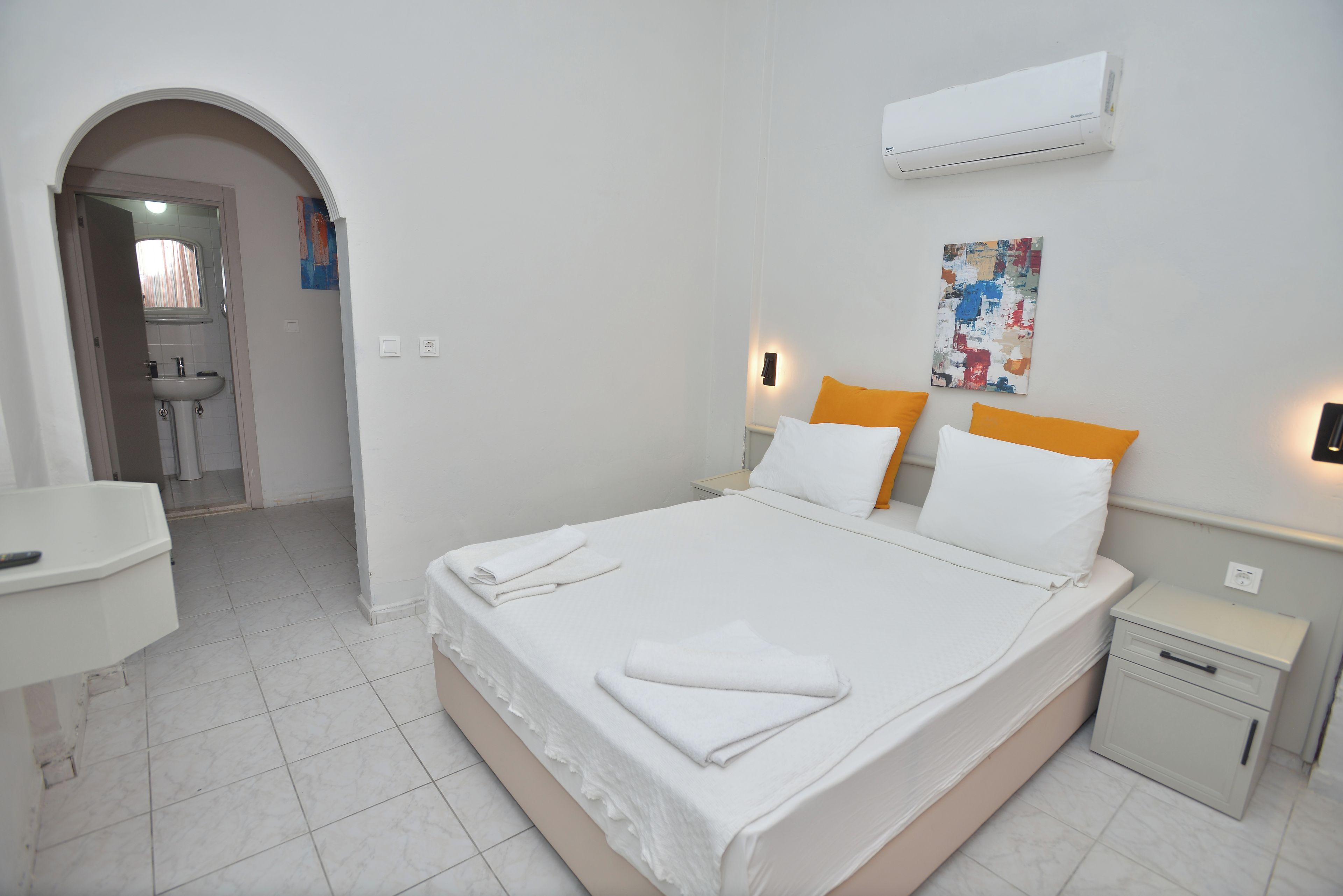 A hotel room with a bed, bedside tables, a desk, and an air conditioning unit.