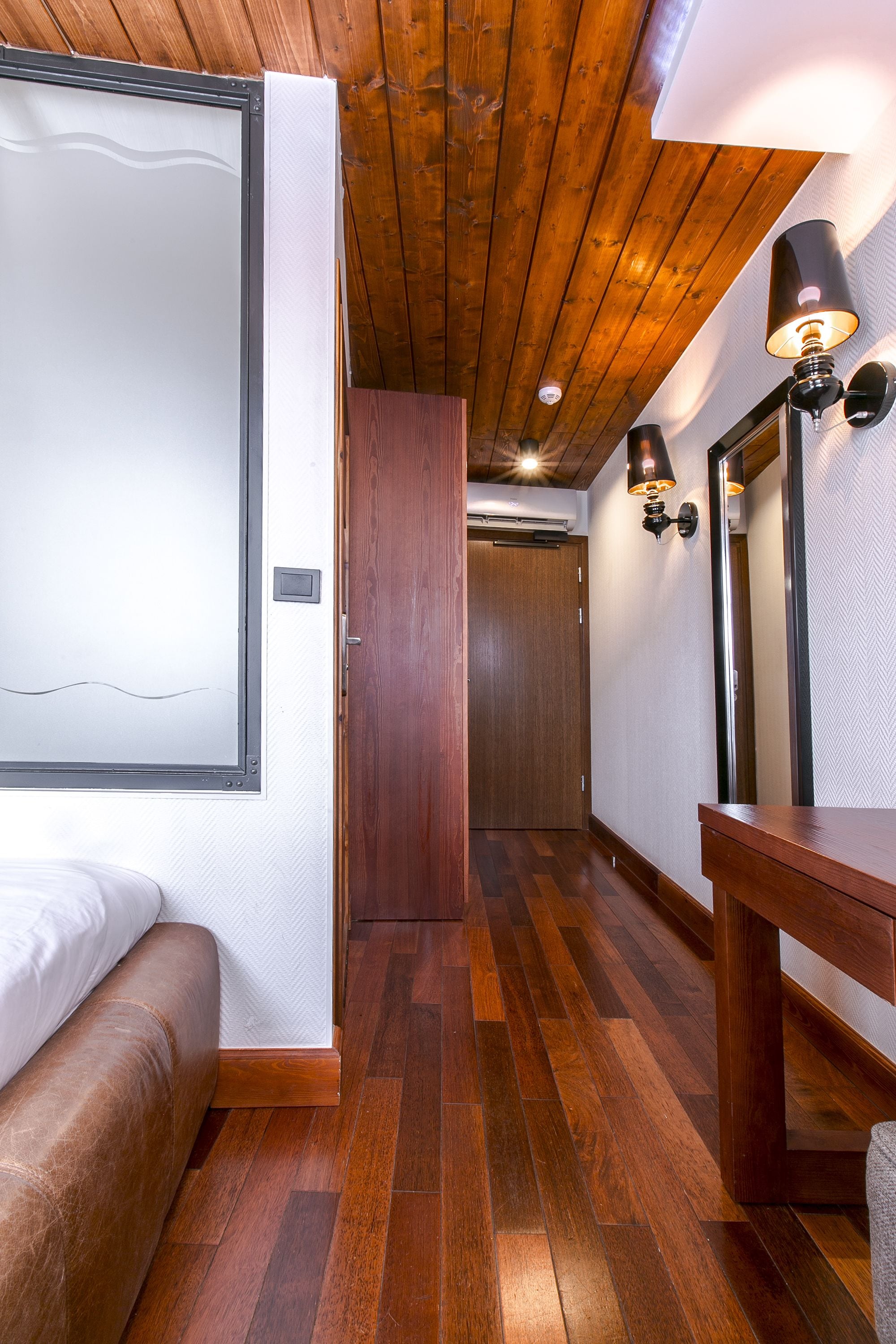 A narrow hallway with wooden flooring, a bed with white bedding, a wooden bench, a wooden table, and a wall-mounted lamp.