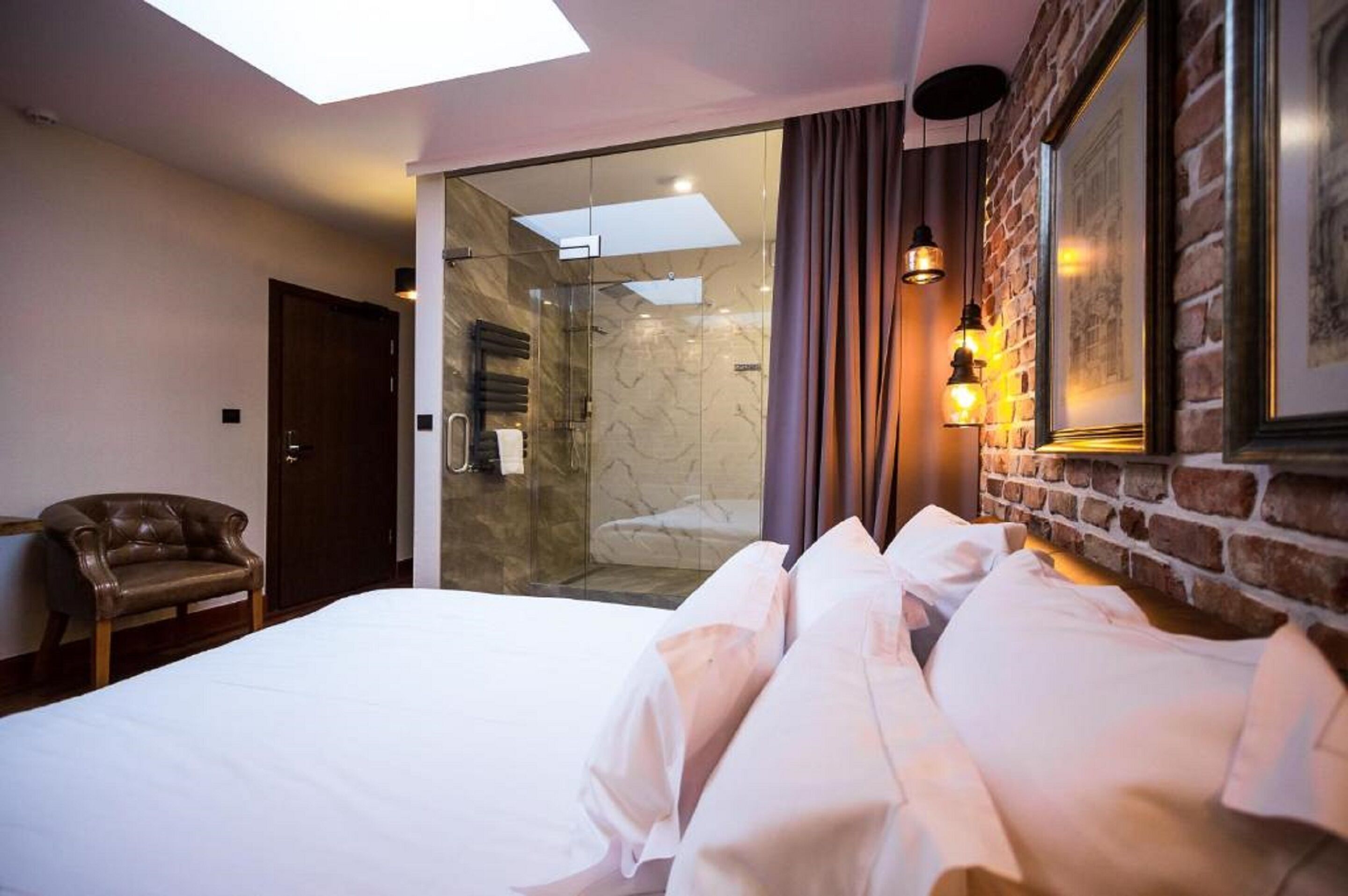 A hotel room with a bed, a leather armchair, a glass shower enclosure, and a brick wall with framed pictures.