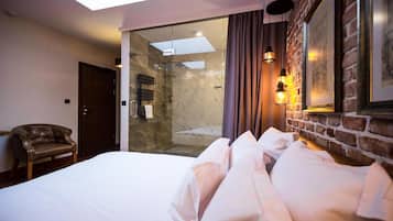 A hotel room with a bed, a leather armchair, a glass shower enclosure, and a brick wall with framed pictures.