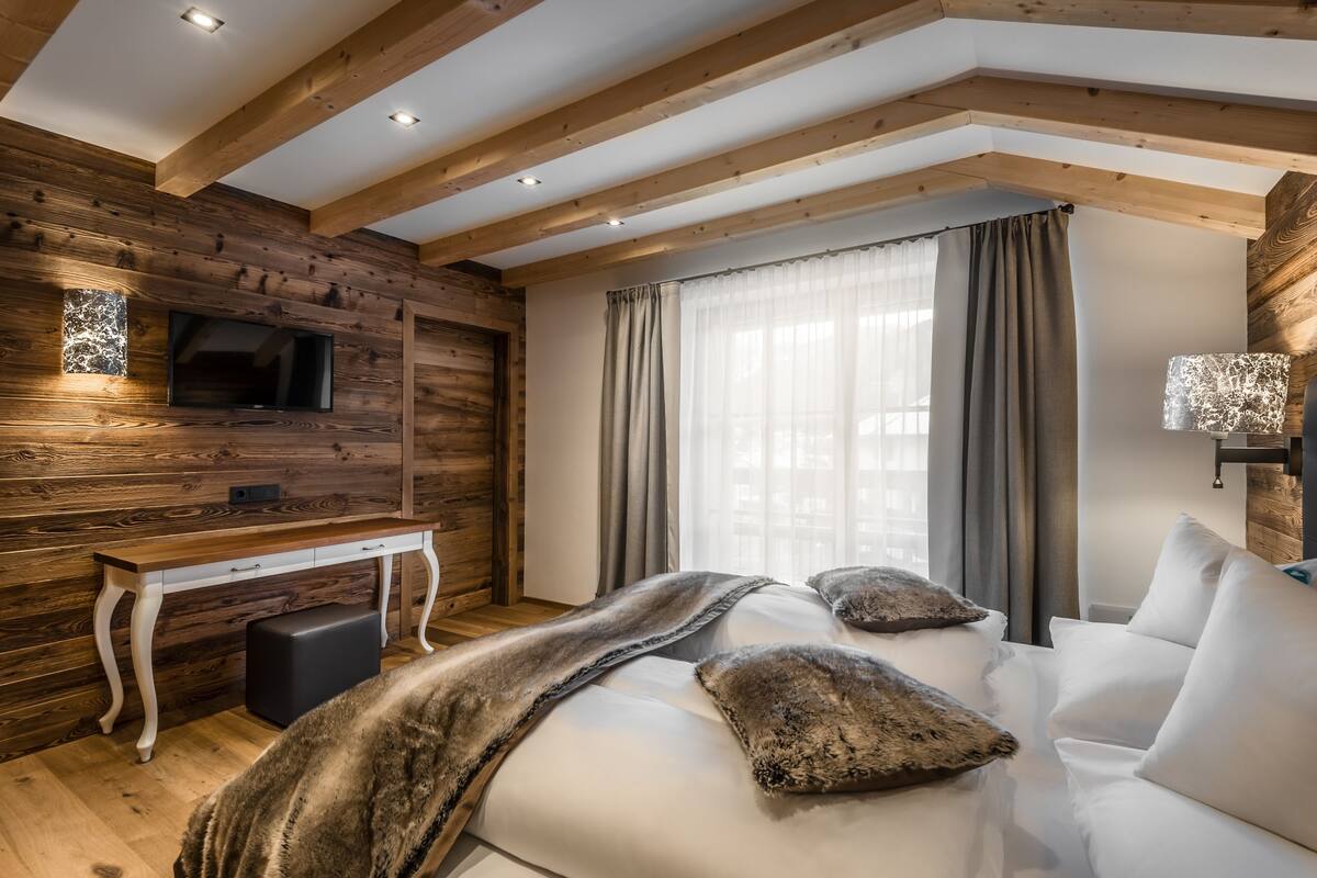Une chambre moderne avec un plafond en bois, un téléviseur à écran plat fixé au mur, un grand lit avec une literie blanche et deux coussins décoratifs, une table de chevet et une fenêtre avec des rideaux.