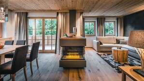 Een moderne woonkamer met een open haard, houten vloeren, een eethoek met stoelen en grote ramen met gordijnen.