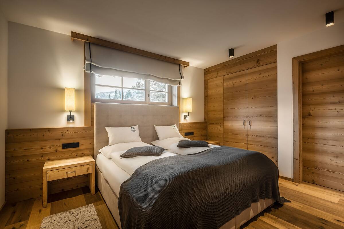 Une chambre à coucher avec une tête de lit en bois, deux lampes de chevet, une fenêtre agrémentée d’un rideau blanc et une armoire en bois.