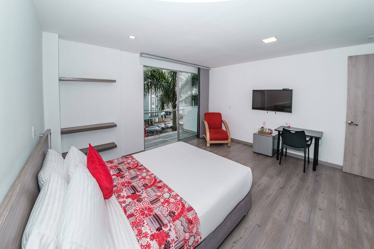A modern hotel room with a large bed, a desk, a chair, a TV, and a sliding glass door to the outside.