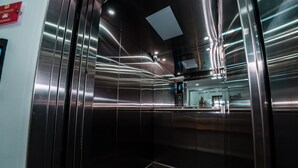 Interior moderno de ascensor con paredes y techo metálicos, una señal de salida de emergencia visible y una pequeña planta en maceta en la pared.